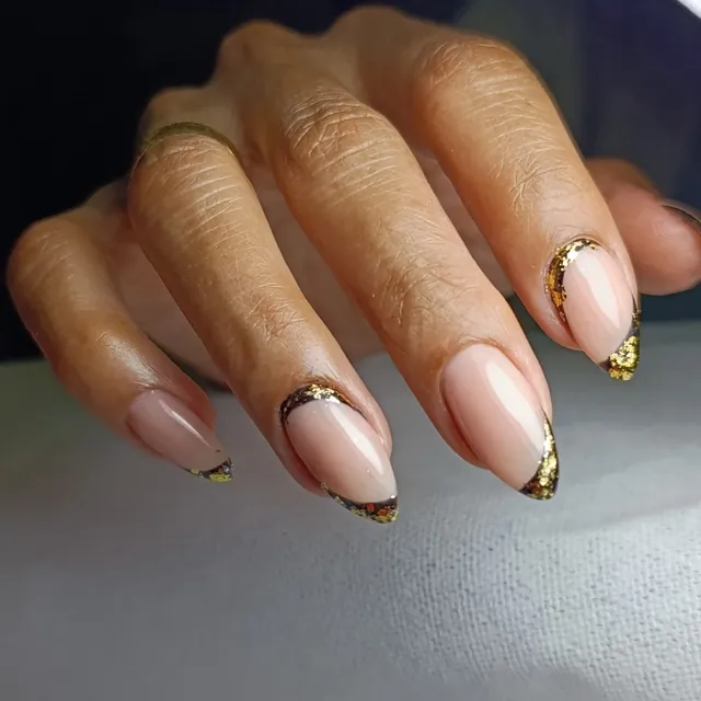 Mano con uñas almendradas en tono nude, puntas negras y láminas de hoja de oro.