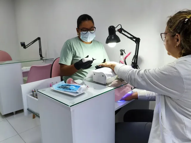 Clienta con la mano bajo la lámpara de curado mientras la manicurista prepara el torno en la mesa de trabajo.