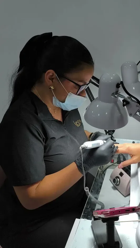 Manicurista concentrada, con tapabocas y guantes, puliendo con torno la uña de una clienta bajo la lámpara.